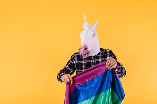 Persona vestida con máscara de unicornio con camisa a cuadros con bandera arco iris lgtbq sobre fondo amarillo Concepto de transexualidad del orgullo gay y derechos lgtbq