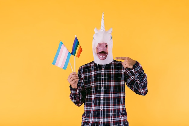 Persona vestida con una máscara de unicornio con una camisa a cuadros que sostiene una bandera de la comunidad transexual y lgtbq sobre un fondo amarillo Concepto de orgullo gay transexualidad género binario y lesbiana