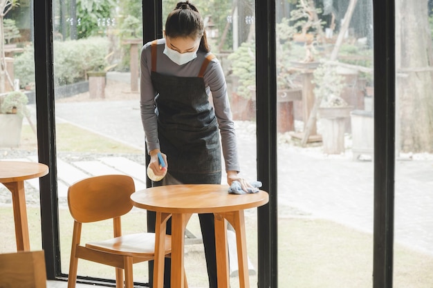 El personal de camarera asiático con mascarilla de protección en la mesa de limpieza del delantal con spray desinfectante protege la infección coronavirus covid19 en la cafetería restaurante concepto de servicio de hotel limpio