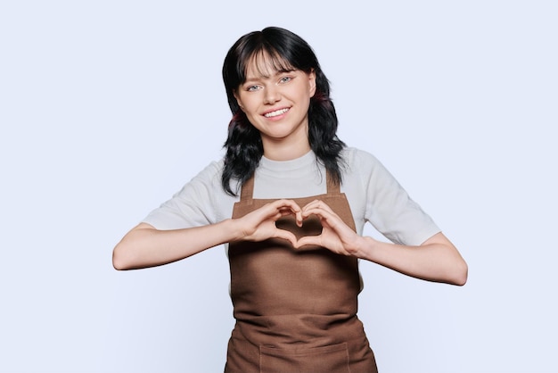 Personal femenino joven sonriente en delantal mostrando gesto de corazón sobre fondo blanco
