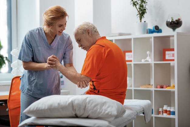 Personal del hospital. Grave anciano sentado en la cama médica mientras es ayudado por una enfermera