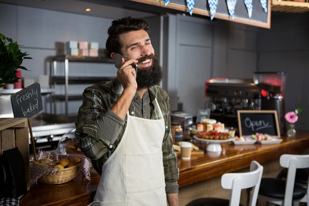 Personal masculino sonriente hablando por teléfono móvil en el mostrador de la cafetería.