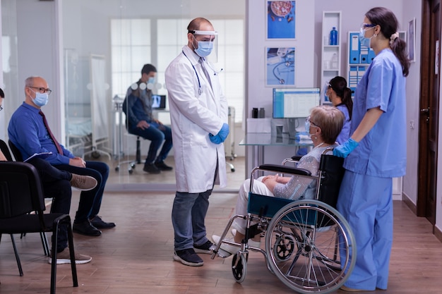 Personal médico con máscara de protección hablando con una mujer discapacitada en el área de recepción del hospital mientras los pacientes esperan el examen