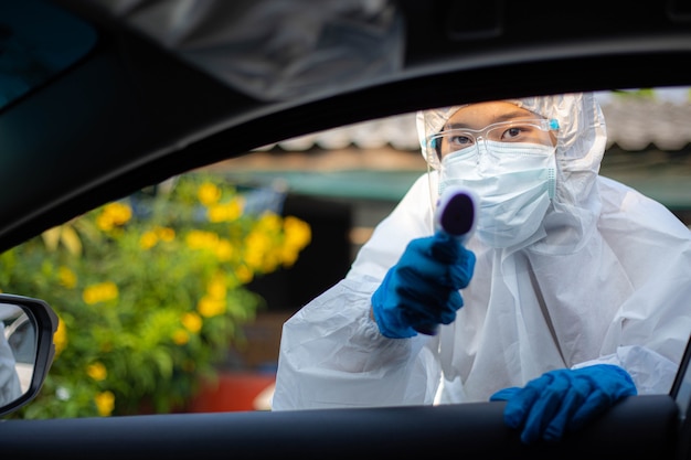 El personal médico con PPE usa una pistola de termómetro infrarrojo para controlar la temperatura corporal en la estación de servicio en el hospital. Nuevo concepto médico y sanitario normal.