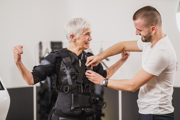 Personal Trainer hilft einer älteren Frau mit Dressing-Anzug für EMS-Training im Fitnessstudio.