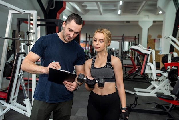 Personal Trainer mit schlanker Klientin, die im Fitnessstudio Bizeps-Curls macht