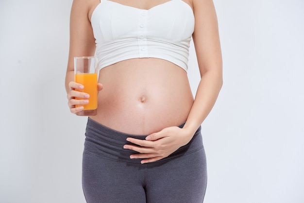 Las personas asiáticas embarazadas de pie en la habitación blanca, ella bebiendo jugo de naranja fresco, usa su mano sosteniendo un vaso