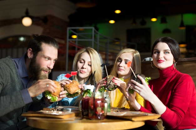 Las personas cenan juntas en una mesa en un café. Amigos felices comen hamburguesas y beben cócteles en el restaurante.