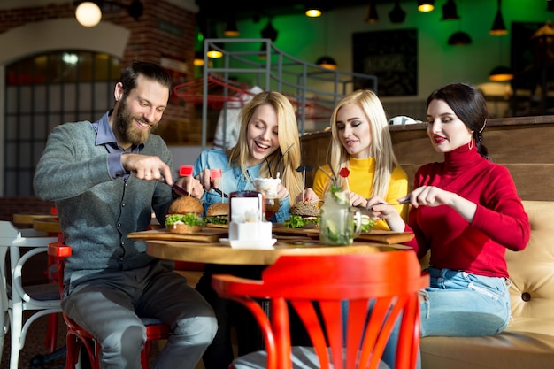 Las personas cenan juntas en una mesa en un café. Amigos felices comen hamburguesas y beben cócteles en el restaurante.