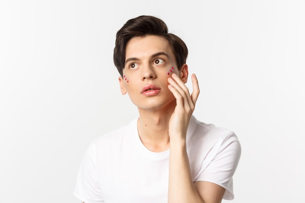 Personas, comunidad lgbtq y concepto de estilo de vida. Hermoso joven gay aplicando brillo debajo de los ojos para la fiesta del orgullo, de pie sobre blanco.