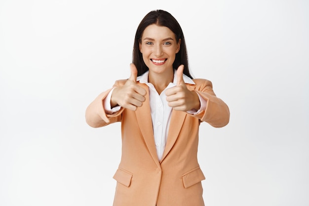 Personas corporativas Sonriente mujer de negocios exitosa mostrando los pulgares hacia arriba con las manos estiradas luciendo complacida satisfecha con un buen resultado de pie en traje sobre fondo blanco