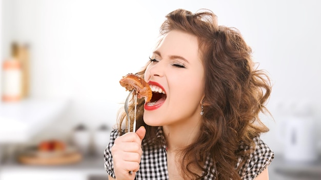 personas, dieta, concepto culinario y alimentario - mujer joven hambrienta comiendo carne en un tenedor sobre el fondo de la cocina