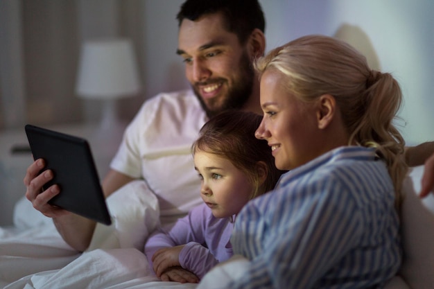 personas, familia y concepto de tecnología - madre feliz, padre y niña pequeña con tableta computadora en la cama por la noche en casa