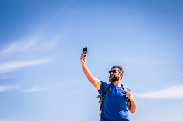 Las personas y el hombre de tecnología de Internet con barba y sunlgasses en busca de señal con un dispositivo de teléfono móvil personas que viajan con mochila por concepto de aventura y cielo azul de vacaciones alternativas