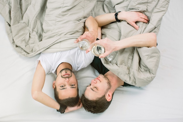 Personas, homosexualidad, familia del mismo sexo, concepto de relaciones homosexuales. Pareja gay masculina abrazándose unos a otros y bebiendo vino blanco mientras está acostado en la cama en la habitación diseñada para loft