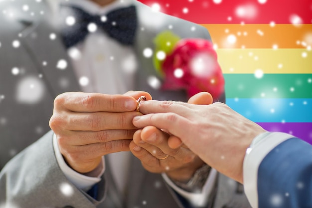 personas, homosexualidad, matrimonio entre personas del mismo sexo y concepto de amor - cierre de las manos de una pareja gay masculina feliz poniendo el anillo de bodas sobre el fondo de la bandera del arco iris y el efecto de la nieve