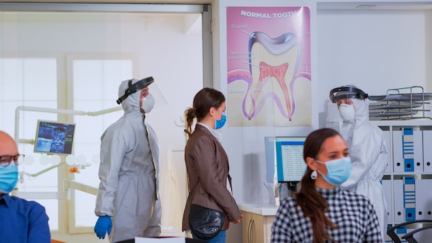 Personas con máscara de protección sentadas en zona de espera respetando la distancia social en nueva recepción normal. Doctor en traje de ppe invitando a paciente a sala de estomatología para examen de dientes durante coronavirus