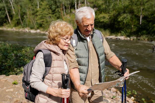 Personas mayores contemporáneas en ropa deportiva mirando el mapa guía mientras intentan encontrar el camino de regreso o continuar su viaje