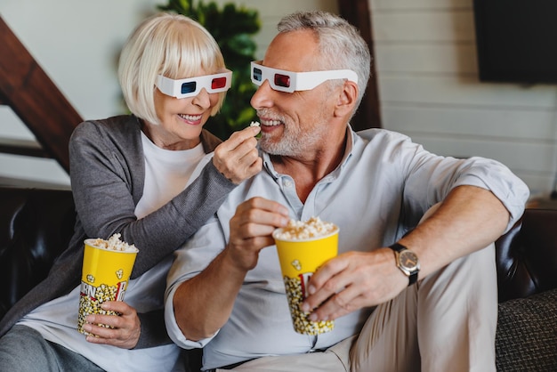 Personas mayores con gafas 3D y palomitas de maíz sentadas en el sofá y viendo películas en casa