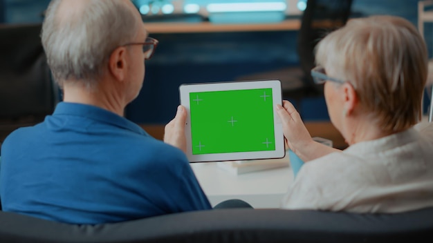 Personas mayores con tableta digital con pantalla verde en la sala de estar. Hombre y mujer jubilados mirando una plantilla simulada aislada con clave de croma en blanco y fondo de espacio de copia en el gadget.