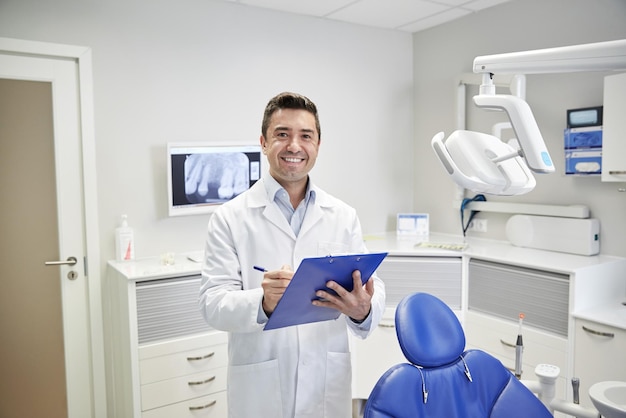 personas, medicina, estomatología y concepto de atención médica - feliz dentista masculino de mediana edad con bata blanca escribiendo en el portapapeles en la oficina de la clínica dental