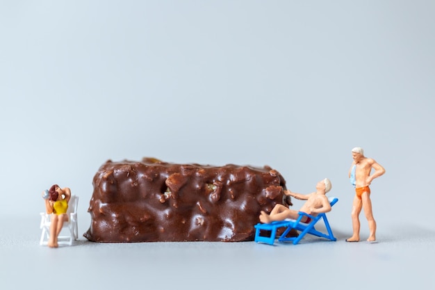 Personas en miniatura con traje de baño relajándose en barras de chocolate sobre fondo gris Concepto del Día Mundial del Chocolate