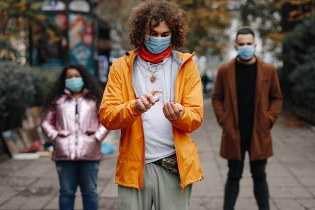 Foto personas multiculturales en máscaras médicas de pie en las calles de la ciudad y con antiséptico. jóvenes amigos que mantienen la distancia social durante la pandemia.