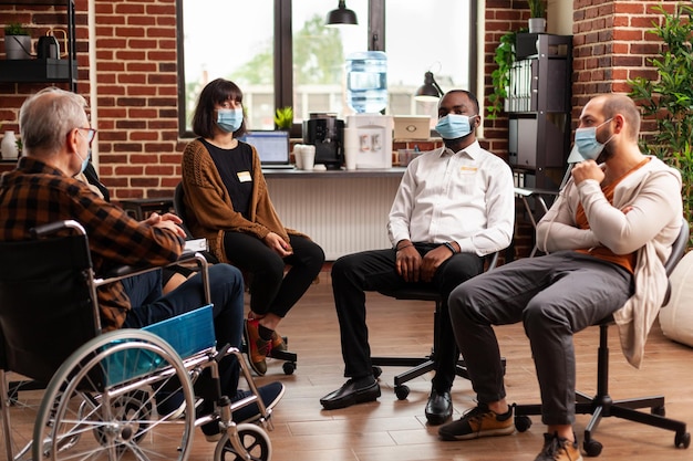 Personas multiétnicas que asisten a sesiones de terapia grupal durante la pandemia de coronavirus. Anciano sentado en silla de ruedas y pacientes en círculo preparándose para una reunión de discusión y asesoramiento.