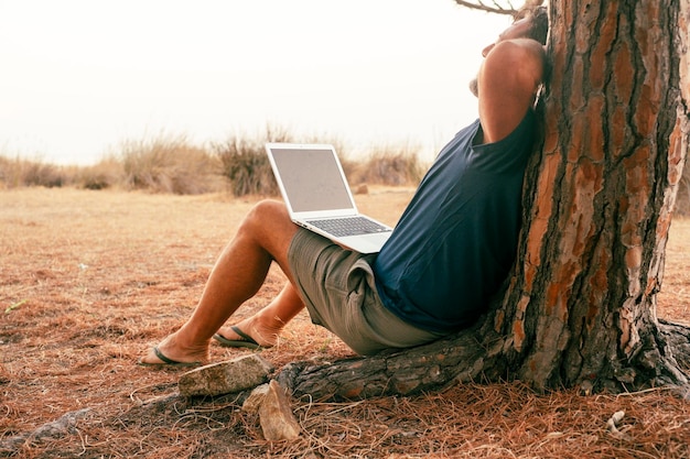 Personas que trabajan al aire libre con computadora y conexión inalámbrica Un hombre estirando su espalda contra un árbol con una computadora portátil en las piernas Concepto de proyecto final y estilo de vida libre de nómada digital Trabajo moderno