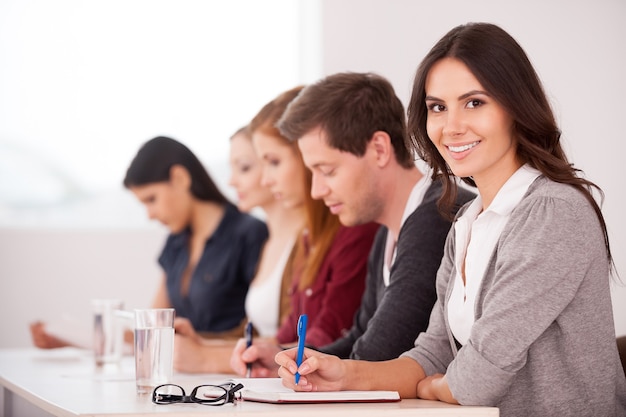 Personas en el seminario. Atractiva mujer joven sonriendo a la cámara mientras está sentado junto con otras personas en la mesa