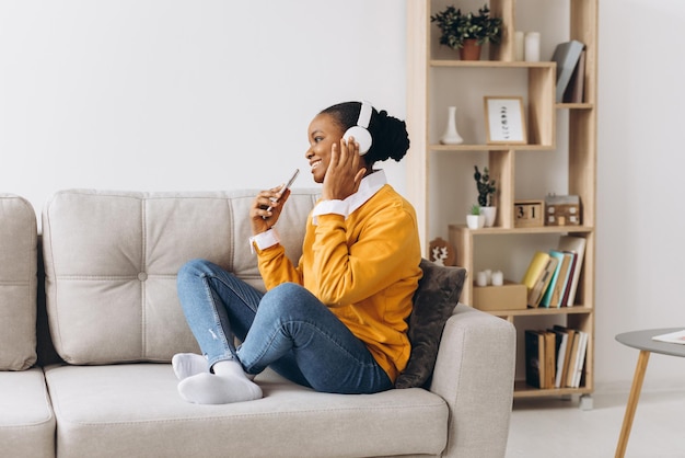 Personas, tecnología y concepto de ocio: feliz joven afroamericana sentada en un sofá con teléfono inteligente y auriculares escuchando música en casa
