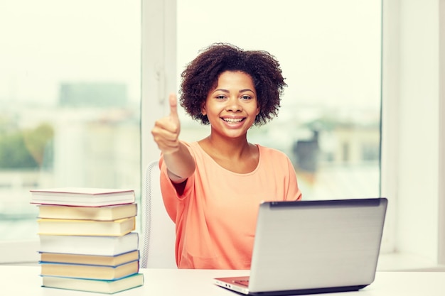 personas, tecnología, gestos y concepto de educación - feliz joven afroamericana sentada en la mesa con computadora portátil y libros mostrando los pulgares hacia arriba en casa