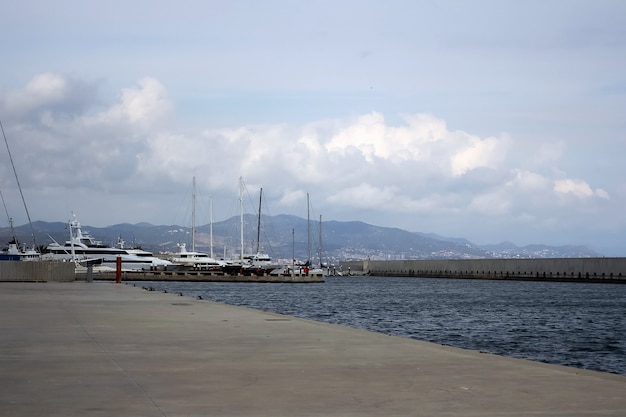 Perspectiva de la orilla del mar con barcos en la superficie rodeados de montañas y nubes