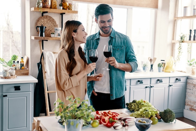 Pertenecen juntos. Hermosa joven pareja cocinando la cena y bebiendo vino mientras está de pie en la cocina de casa