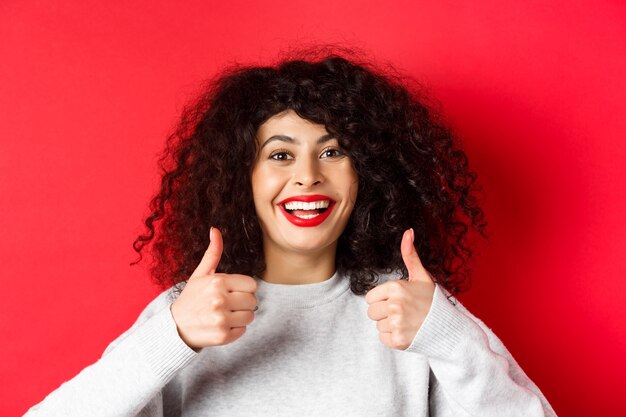 Perto da alegre mulher caucasiana, mostrando os polegares em aprovação, sorrindo satisfeito, gosto e elogio algo bom, de pé sobre fundo vermelho.