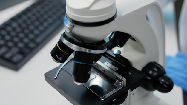 Perto da bandeja de amostra do microscópio científico em laboratório, usada pela mão do médico com luvas para pesquisa. Ferramenta de química óptica com lente e lupa para estudo de DNA e bactérias