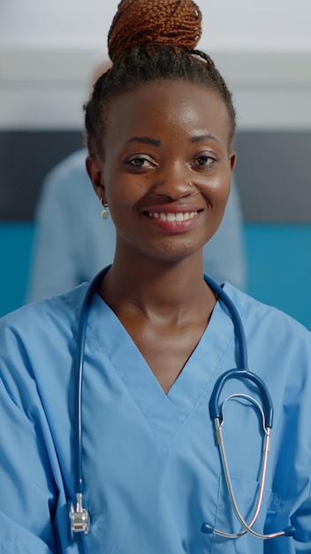 Perto da enfermeira afro-americana, usando o computador laptop na mesa branca na clínica de saúde. Assistente médico negro digitando no dispositivo enquanto olha para a câmera com estetoscópio e uniforme