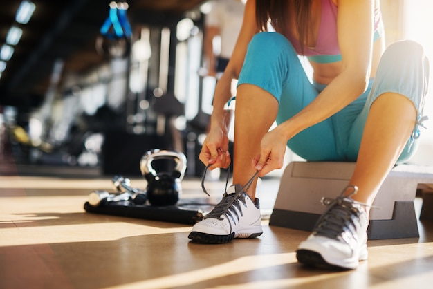 Perto da garota de fitness magro jovem sentado no degrau e amarrando o cadarço no ginásio.