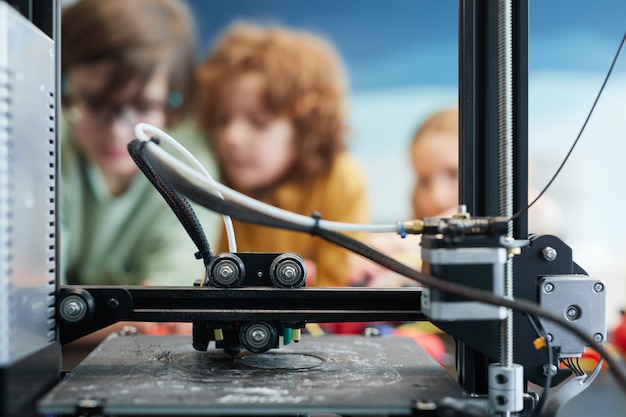 Perto da impressora 3D fazendo modelos de plástico durante a aula de engenharia e robótica na escola com crianças no fundo, copie o espaço