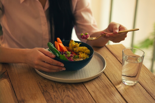 Perto da mesa do café e um copo de água com uma tigela de legumes. Mão de mulher comendo comida com um garfo