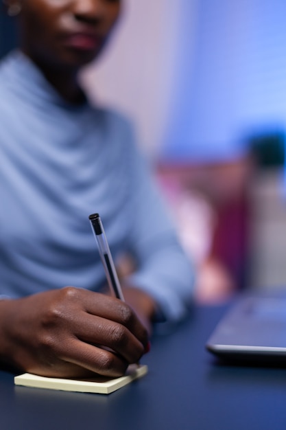 Perto da mulher africana tomando notas, sentado na mesa à noite para o trabalho. freelancer negro respeitando prazo de estudo tarde da noite.