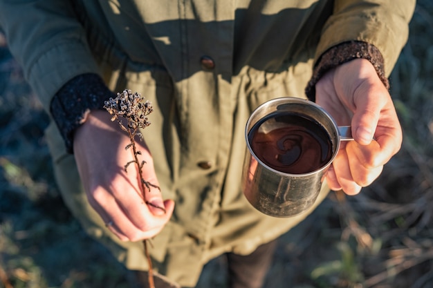 Perto da mulher com uma xícara de café e flores em uma bela luz do sol. Beleza no conceito de natureza: mãos femininas segurando um copo de bebida quente e uma flor seca do campo.