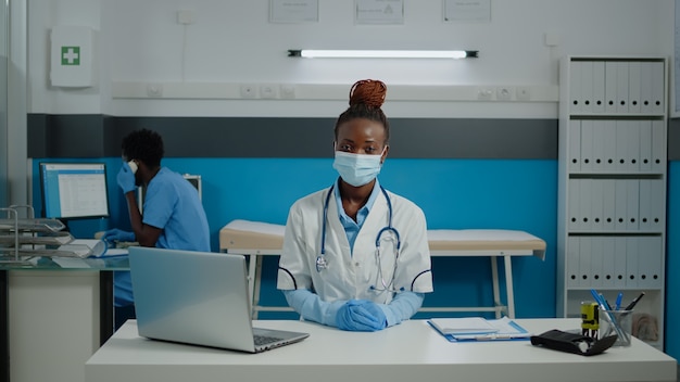 Perto da mulher que trabalha como médica no gabinete médico na instalação. retrato de jovem médico usando máscara facial e sentado à mesa com laptop, documentos, arquivos e papéis de check-up