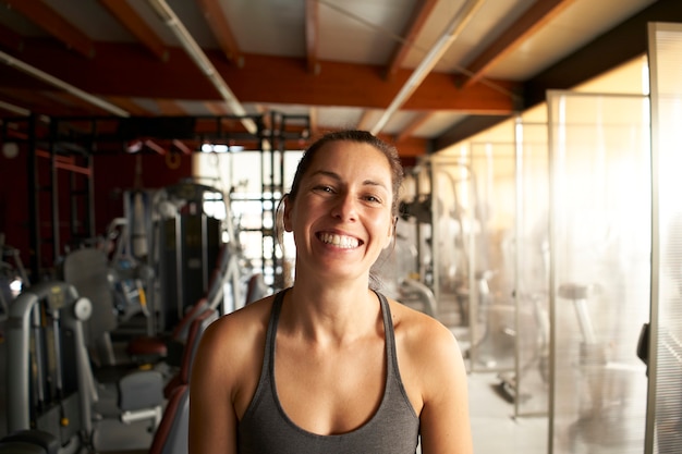 Perto da mulher sorridente jovem fitness em pé no ginásio, olhando a câmera.