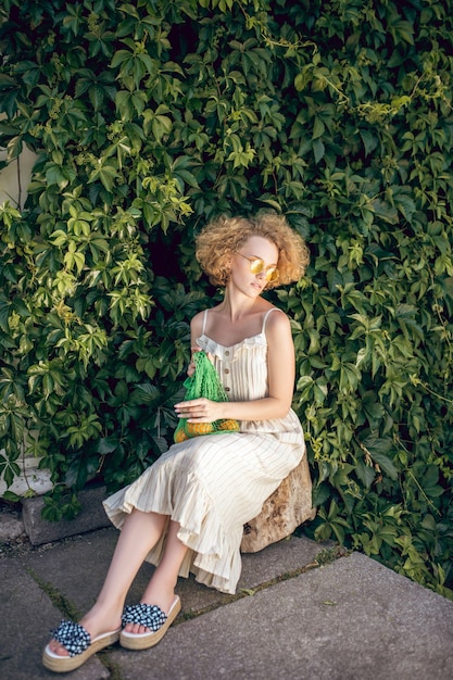 Perto da Natureza. Mulher bonita em um vestido longo branco e óculos escuros segurando uma bolsa de malha verde nas mãos