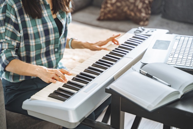 Foto perto das mãos de uma jovem irreconhecível, tocando piano elétrico, ensinando remotamente, usando o laptop enquanto trabalhava em casa. conceito de educação e lazer online.