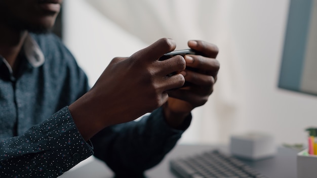 Perto das mãos do homem negro jogando videogames para celular na Internet online em seu telefone no lazer doméstico ...