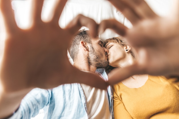 Perto de casal apaixonado beijando fazendo formato de coração com as mãos deitado na cama.