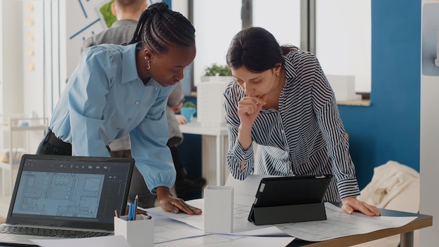 Perto de diversas mulheres, analisando o plano de plantas com tablet digital, trabalhando em equipe para o projeto. arquitetos trabalhando no layout e estrutura de construção para desenvolvimento urbano.