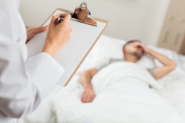 Perto de médico escrevendo em um prontuário médico com paciente deitado em uma cama de hospital